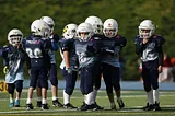 A group of nine maybe six- to eight-year-old football players in full football gear are in a huddle on a turf field ready for their coach to call a play.