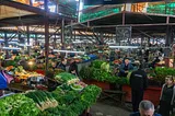 A view of a covered market hall in Kutaisi, Georgia. There are displays of fresh veggies along with vendors and shoppers.