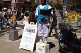 A man, dressed elaborately as an 18th century town crier, stands on a soap box next to a sign reading, “Town crier for your Tweets.”