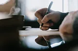 A side POV of hands holding a pen about to write on paper. Further back is a coffee cup with steam coming out of it.