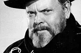 A black and white photograph of Orson Welles. He is wearing a thick black coat and scarf and a wide-brimmed black hat. He is older, late 50s, with a greying beard. He is looking directly at the camera with a smouldering expression. The photo is signed by the actor.