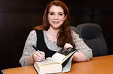 Author Stephenie Meyer sitting at a desk and signing a copy of her book.