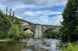 An ancient stone bridge with high arches over a tranquil river that you see in the foreground. It’s a sunny day, a blue sky with some smaller clouds. Left and right of the river are green trees.