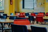 A vibrant classroom setting with rows of empty desks and colorful chairs in shades of red and blue. The arrangement suggests readiness and potential for learning, while the absence of students invites reflection on the dynamics and leadership that shape educational spaces. The polished wooden floors and natural light streaming in create a warm yet thought-provoking atmosphere, emphasizing the contrast between the physical environment and the unseen forces of human behavior that influence a child