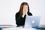 Dark-haired white woman sits in front of Macbook, eyeglasses on the desk, hands pressed over her face, looking stressed or exhausted