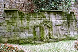 Carved in relief on an old stone wall is a plaintive female figure, arms outreached, eyes skyward, who can be clearly seen. Less visible are the much shallower reliefs, the carvings of people, mostly men with mustaches, who seem to hover around her.