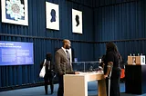 Exhibition space with white artworks on blue walls, display pedestals, and engaged gallery visitors.