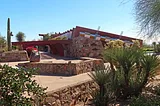 Desert brush in the foreground with a low rock wall between the garden and a large beige concrete pad. There is a low rocks ledge in a square shape with a large rock in the corner, a saguaro cactus is in the background beside a stone faced one-level sloped-roof building with a red metal portico