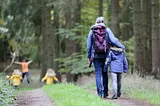 Woman in gray coat and jeans with a purple backpack walking on a forested path with her arm around a child in a blue coat. In the background, three other children walking and running ahead.