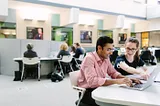 Two students sit around a computer in a library