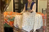 A.D. Herzel, Korean American Adoptee woman in front of a mirror in a white and blue Traditional Afghan dress, with gold embroidered trim.