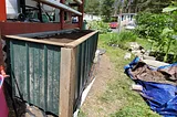 A raised garden bed with wood trim and green aluminum siding in a yard in front of a deck painted a red-brown color. The lawn in front of the raised bed is torn up and on the right are blue tarps with dirt piled on. Behind the tarps is a raised bed made from concrete bricks with tall sunflower plants.