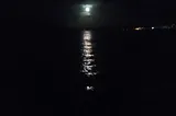 Photo of the moon coming out from the clouds and shining over a bay —  leaving a trail of light on the dark water.