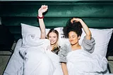 Two women lie under covers on a white bed with funny expressions and arms raised.