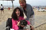 A blond man leaning over a long-haired woman in a wheelchair holding a little dog. They are on the beach.