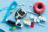 A collection of items associated with diabetes including a blue ribbon, glucose monitor, candy, sugar cubes, insulin syringe and a pink donut with sprinkles.