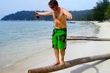 A tanned male in green board shorts balancing on a fallen palm trunk on a white sandy beach.