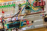 Photograph of electronics prototyping boards with many wires, an LED display that says ‘COdE’ and, at bottom left, a chip in a socket.