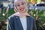 A woman in her 20s wearing a pink hijab is smiling at the camera, shown shoulders up. Behind her is a city park and buildings.