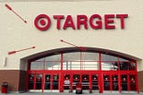 A Target store entrance is shown with the brand name prominently displayed above red double doors. Several red arrows have been added to the image, pointing at the bullseye logo. The arrows emphasize the play on the word “Target.”