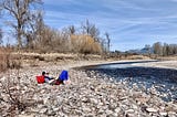 The author reading a book on a pebble beach next to a channel of the Clark Fork River in Missoula, MT