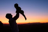 Color photo. In the foreground, a father lifts his baby son up above his head. In the background is a stunning sunset.