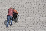 A woman pushes an elderly woman in a wheelchair across a sidewalk.