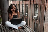 Smiling Black woman sitting on a terrace with her laptop, on a phone call.