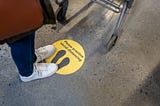 A photo of a person standing on a sticker that says “practice social distancing” at a grocery store.