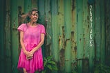 Photograph of an older woman wearing a pink dress and standing in front of a rusted shipping container