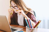 Female student bites on a pencil while looking at her laptop screen.