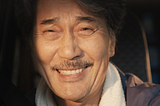 A close-up of Hirayama smiling and half-crying. He has a large almost pained grin, his lips are pursed and you can see his teeth. He has a small moustache. You can see the car headrest behind his head. The sun is shinning into his face. You can only see the top part of his blue overalls. His overalls have a white collar.