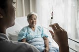 A photo of a nurse holding up a syringe with an elderly woman sitting in a hospital bed in the background.