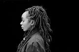 Black and white image of a Black woman with dreadlocks against a black ground.