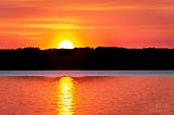 The sun rises over (and reflects off of) a calm lake in upstate New York.