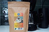 A paper bag of coffee on a kitchen counter in front of a coffee maker and a coffee grinder. The label on the bag says: ‘Swiss water process. Koffeinfri. Masha Estate. Etiopia. 250g hele bønner. Koffeinfri kaffe med god, kremet fylde, frisk syrlighet. Hint av jordbær, honning og mango i smaken.’