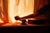 Amber light glows through sheer curtains, backlighting the table where a woman reaches for a cup sitting neatly in its saucer.