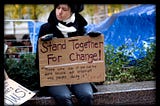 A woman in jeans and a black coat and black and white scarf sitting on a concrete bench at Occupy Wall Street, with a blue tarp in the background. She is holding a sign that reads “Stand together for change! Those say it can not be done should not interrupt the people doing it. Join us!”