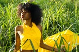A woman of African descent with mid-length curly hair poses in a flowing yellow dress with a trail. Her face is exposed to natural lighting from the sun though her eyes are closed, she’s posing in a field of long, green grass. it’s visible both in the foreground, and the background.