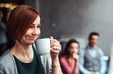 A woman smirks as she holds a coffee mug up to her face in front of 2 coworkers.