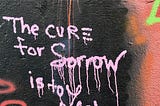 Light pink colored spray-painted graffiti writing on a wall of mainly black with some other colorings wall stating “The cure for sorrow is to learn something”.