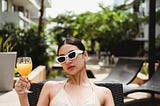 Confident young ethnic lady drinking juice lying on sunbed at poolside