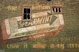 An old, faded ad for Wrigley’s spearmint gum painted on a brick wall pierced by two broken windows.