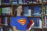 Rebecca in a “Library Student Organization” superman-themed t-shirt holding a book in front of the stacks