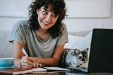 Woman smiling looking at her laptop while writing in a notebook with a cat beside her focused on the laptop.