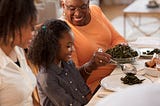 A smiling grandmother serves vegetables to her granddaughter at the dining table.