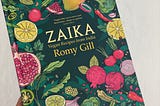 A hand holding the book “Zaika” by Romy Gill, which has illustrations of various vegetables and fruits on a dark green cover