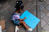 A young Black girl sits on the ground with a bright blue sign that says “Black Kids Matter.”