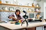 Adult woman preparing a meal and chopping fresh ingredients in front of a video camera on a tripod.