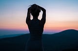 This photograph captures the silhouette of a woman standing against a twilight sky. She is seen from behind, wearing a fitted sleeveless dress with horizontal stripes. Her arms are raised as she gathers her hair into a ponytail. The background showcases rolling hills fading into the horizon, bathed in the soft hues of a sunset — shades of pink, orange, and deepening blue. The scene evokes a quiet, contemplative mood.